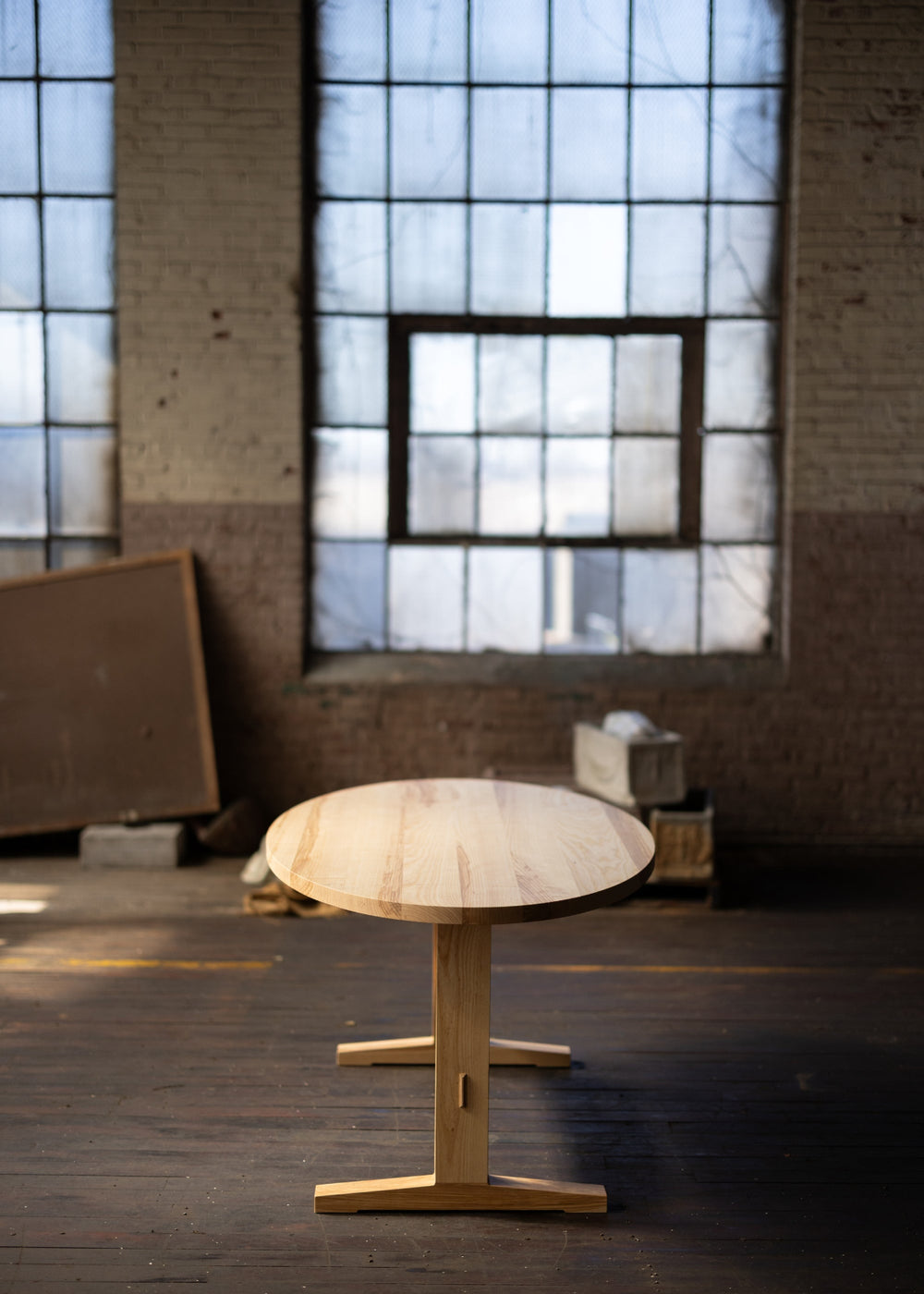 Wooden shaker style oval table in a room with large windows and exposed brick walls.