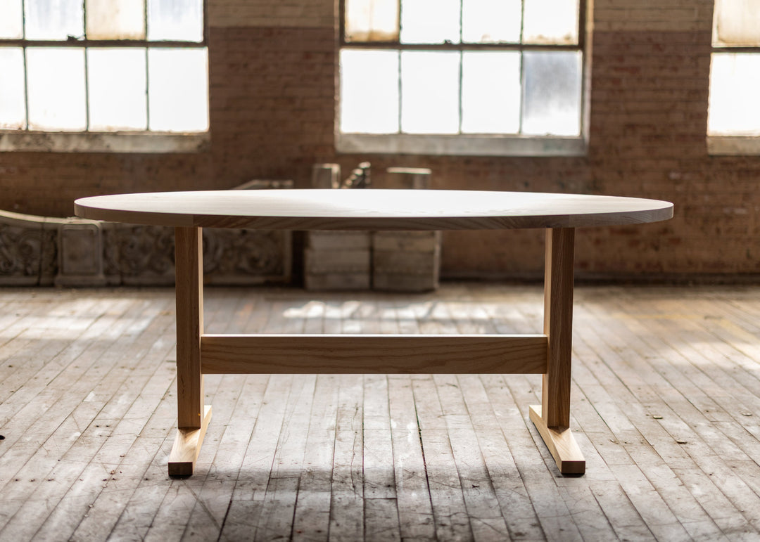 Oval ash wooden pedestal shaker style table in a room with large windows and brick walls.
