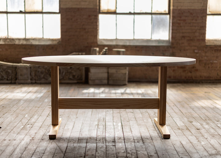 Oval ash wooden pedestal shaker style table in a room with large windows and brick walls.